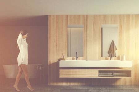 Young woman with dark hair walking in modern bathroom with white and wooden walls, comfortable double sink and bathtub in background. Toned image double exposureの写真素材