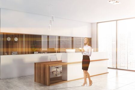 Thoughtful young blonde businesswoman standing in modern panoramic office with white walls, tiled floor and comfortable white and wooden reception desk. Toned imageの写真素材