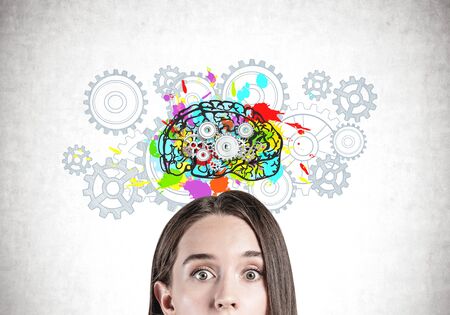 Head of astonished young woman standing near concrete wall with colorful brain sketch with gears drawn on it. Concept of creative thinking and brainstormingの写真素材