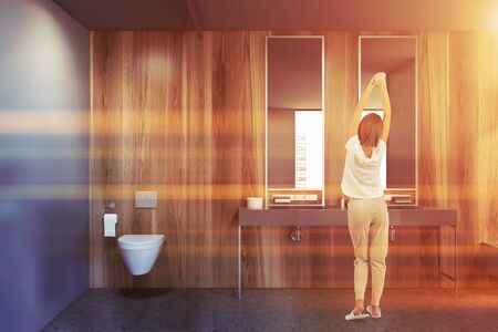 Rear view of young woman in pajamas standing in modern bathroom with gray and wooden walls, concrete floor, double sink and toilet. Toned imageの写真素材