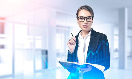 Beautiful young businesswoman with glasses holding notebook and pencil and standing in blurry office with double exposure of financial chart. Concept of planning and stock market. Toned imageの写真素材