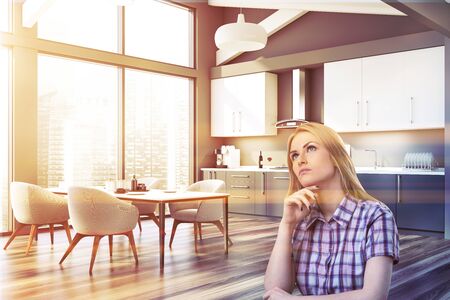 Thoughtful blonde woman sitting in modern kitchen with gray walls, wooden floor, grey countertops and wooden table with white armchairs. Toned imageの写真素材