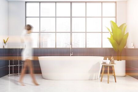 Blurry young woman walking in stylish bathroom with white and blue tiled walls, concrete floor, big window and comfortable bathtub. Concept of spa. Toned imageの写真素材
