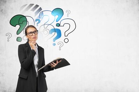 Pensive young businesswoman in glasses holding notebook near concrete wall with colorful question marks drawn on it. Concept of choice and education. Mock upの写真素材