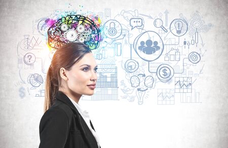 Side view of confident and successful young businesswoman with long fair hair standing near concrete wall with colorful brain sketch and business icons drawn on it. Concept of planningの写真素材