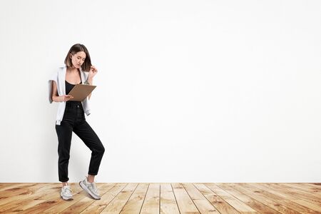 Portrait of beautiful European college girl in casual clothes reading document on clipboard and touching her hair. Concrete wall background. Concept of education. Mock upの写真素材