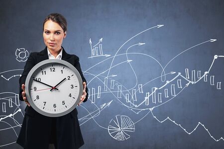 Beautiful young European businesswoman showing big office clock standing near blackboard with graphs...
