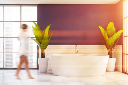 Blurry young woman walking in luxury bathroom with blue and white brick walls, tiled floor and comfortable bathtub with two big potted plants near it. Toned imageの写真素材