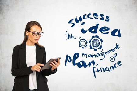 Serious young businesswoman in glasses using tablet near concrete wall with creative business idea sketch drawn on concrete wall. Concept of success, technology and planningの写真素材