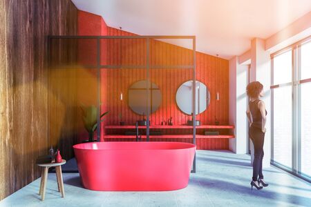 Attractive young African American woman standing in luxury panoramic bathroom with white, red and wooden walls, comfortable red bathtub and double sink. Blurry cityscape. Toned imageの写真素材