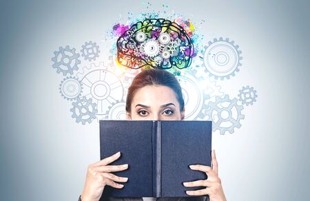 Portrait of young woman covering her face with black book standing near gray wall with colorful...