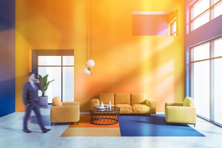 Blurry young man walking in bright living room with blue and orange walls, concrete floor, comfortable orange sofa and armchairs near round coffee table. Toned imageの写真素材