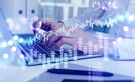Hands of businesswoman typing on laptop in blurry office with double exposure of financial charts...