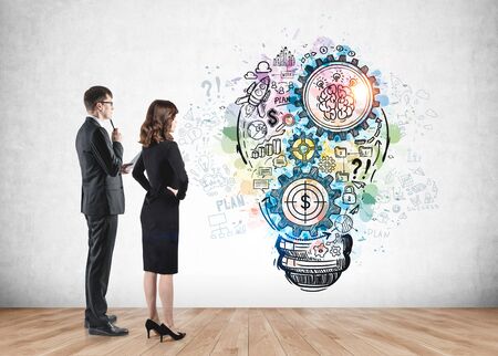Thoughtful businessman and confident businesswoman looking at colorful business idea sketch drawn in concrete wall room. Concept of business strategy and teamworkの写真素材