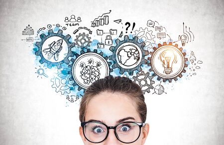 Close up portrait of astonished teen girl in glasses standing near concrete wall with creative gears sketch. Concept of brainstorming and teamworkの写真素材