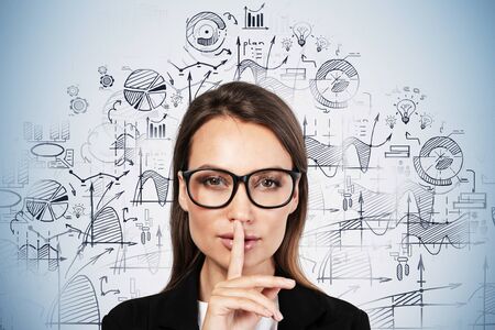 Beautiful young European businesswoman in glasses making hush sign near blue wall with blurry...