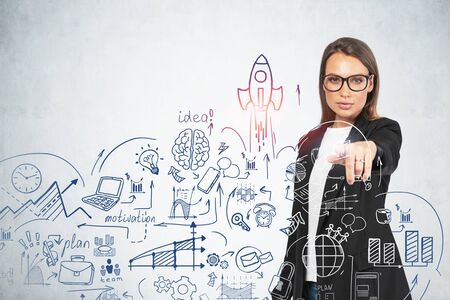 Beautiful young European businesswoman in glasses pointing at creative startup sketch near concrete wall. Concept of new project launch and entrepreneurship. Double exposureの写真素材