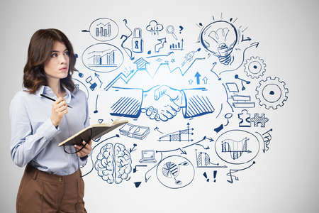 Pensive young European businesswoman with notebook standing near gray wall with creative business partnership sketch drawn on it. Concept of deal and teamworkの写真素材