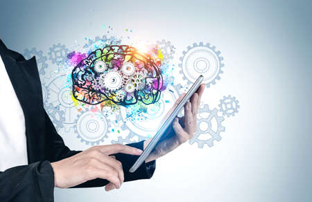 Hands of businesswoman using tablet computer near gray wall with colorful brain sketch drawn on it. Concept of brainstorming and artificial intelligenceの写真素材