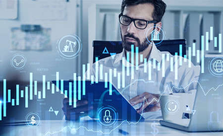 Serious young bearded trader in glasses working in his blurry office with double exposure of digital...