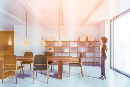 Beautiful young African American woman standing in luxury dining room with white and brown walls, massive table with gray chairs and shelves. Blurry cityscape. Toned imageの写真素材