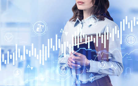 Unrecognizable young businesswoman with notebooks standing in blurry office with double exposure of financial graphs. Concept of trading and market analysis. Toned imageの写真素材