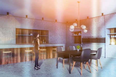 Beautiful young African American woman standing in spacious loft kitchen with stone and wooden walls, bar counter with stools and black dining table. Toned imageの写真素材