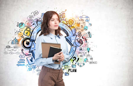 Serious young European businesswoman with notebooks standing near concrete wall with colorful business goal sketch drawn on it. Concept of planning and education. Mock upの写真素材