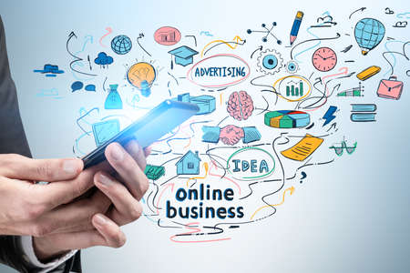 Hands of businessman holding smartphone near gray wall with colorful online business sketch. Concept of e commerceの写真素材