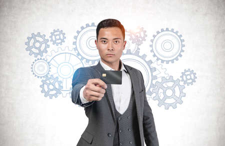 Serious young Asian businessman showing credit card standing near concrete wall with gears sketch drawn on it. Concept of brainstorming and teamworkの写真素材