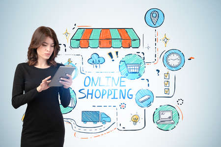 Serious young woman in black dress using tablet near blue wall with colorful online shopping sketch. Concept of e commerceの写真素材