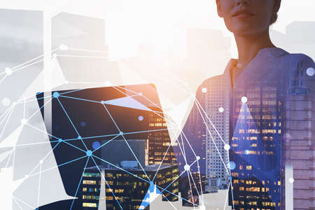 Unrecognizable young businesswoman using laptop in blurry night city with double exposure of network...