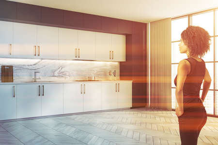 Beautiful young African American woman standing in minimalistic kitchen with white walls, wooden...
