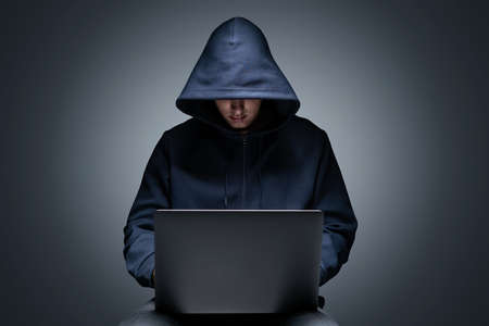 Portrait of unrecognizable young hacker in black hoodie using his laptop over gray wall background....