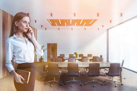 Blonde young businesswoman talking on smartphone in meeting room with white and wooden walls and two...