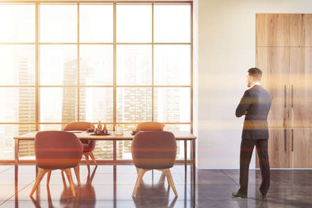 Young man in suit standing in panoramic dining room with white walls, concrete floor and dining...