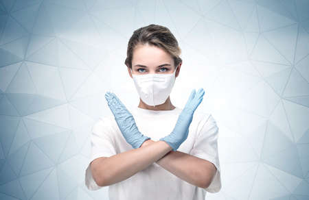 Portrait of serious young European woman in protective mask and glasses showing stop coronavirus sign near gray wall.の写真素材