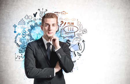 Pensive young businessman standing near concrete wall with business goal sketch drawn on it. Mock up
