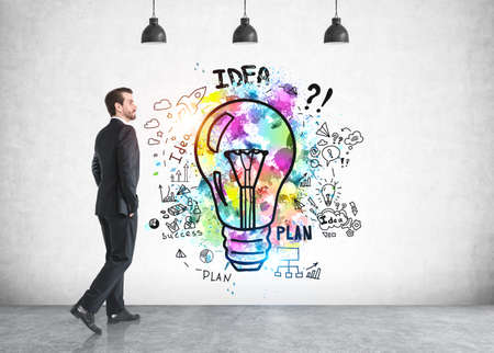 Side view of young businessman walking near concrete wall with bright business idea sketch drawn on itの写真素材