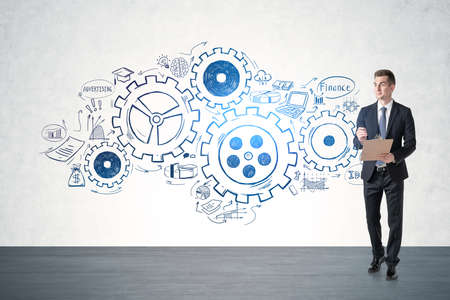 Young businessman with clipboard walking in concrete room with gears sketch drawn on the wall. Concept of teamworkの写真素材