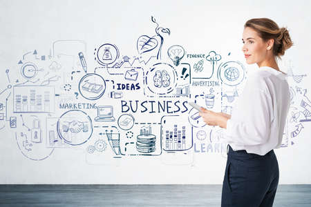 Young European businesswoman standing with tablet near concrete wall with creative business sketch drawn on itの写真素材