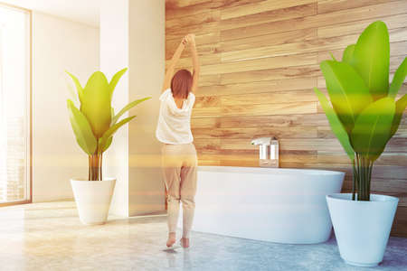 Young woman standing in modern bathroom with white and wooden walls, concrete floor and comfortable...
