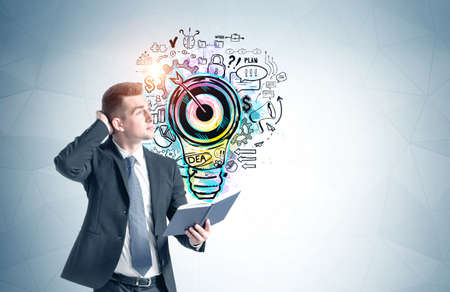 Pensive young businessman with notebook standing near blue wall with colorful business goal sketch...