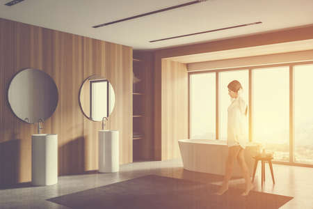Blurry young woman walking in wooden bathroom corner with white tub and double sink with two round mirrors. Toned image double exposureの写真素材