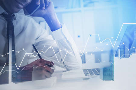 Unrecognizable young African American businessman talking on smartphone in blurry office with double exposure of financial graphs. Toned imageの写真素材