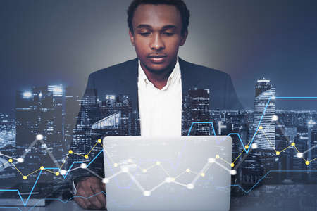 Portrait of serious young African American businessman using laptop in blurry city with double...