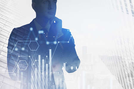 Pensive young businessman standing in blurry city with double exposure of digital charts. Toned...