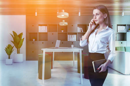 Young woman talking on smartphone in panoramic green living room with wooden walls, sofa, table and bookcase. Toned imageの写真素材