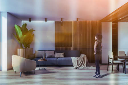 Young African American woman standing in modern living room with white and wooden living room with gray sofa and armchair. Toned imageの写真素材