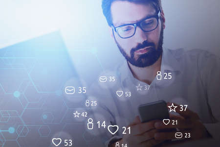 Young businessman in glasses using computer and smarpthone in blurry office with double exposure of social media interface. Toned imageの写真素材